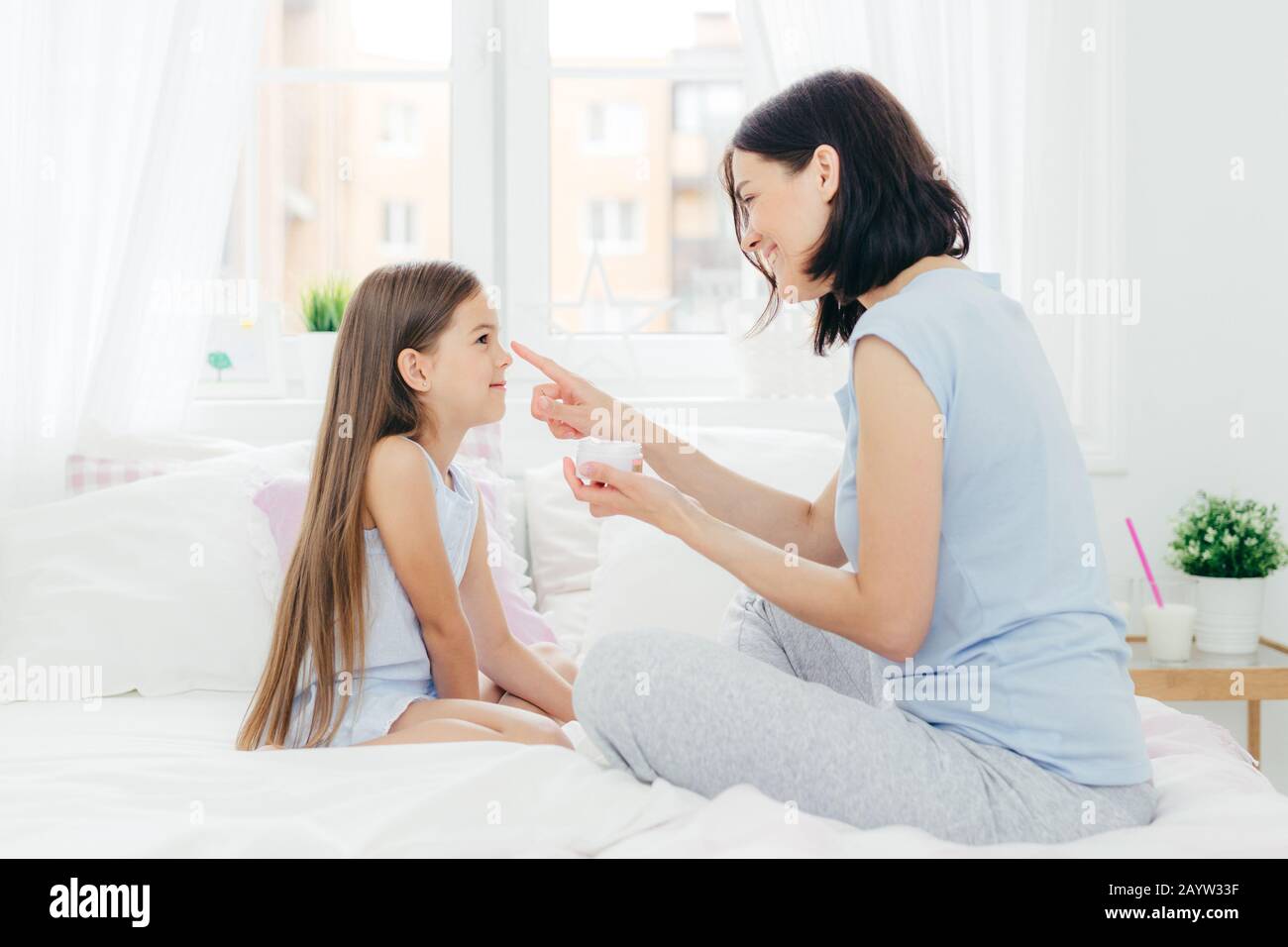 Affectionate mother applies cream on daughter`s nose, sits together at bed, takes cake of healthy skin and good appearance, have beauty procedure at h Stock Photo
